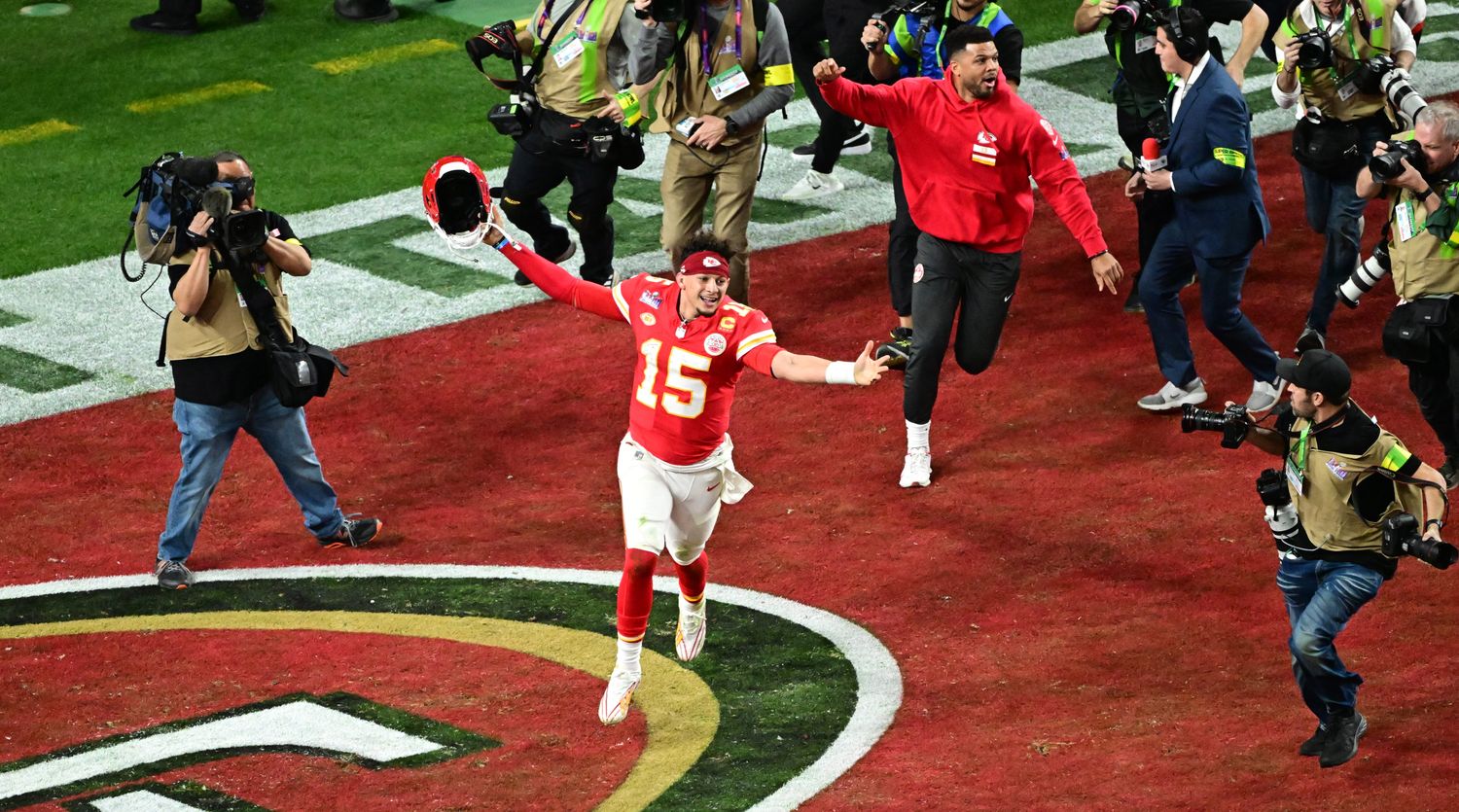 Patrick Mahomes runs with his arms out holding his helmet in the end zone as camera men run behind him